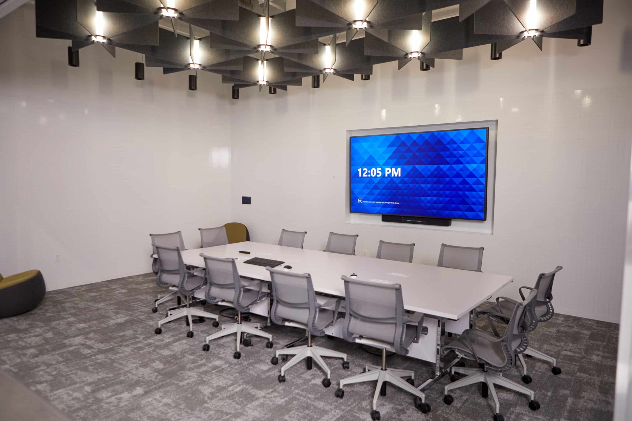 Modern conference room: rectangular table, 10 gray swivel chairs, large screen shows 12:05 PM, geometric acoustic panels, gray carpet.
