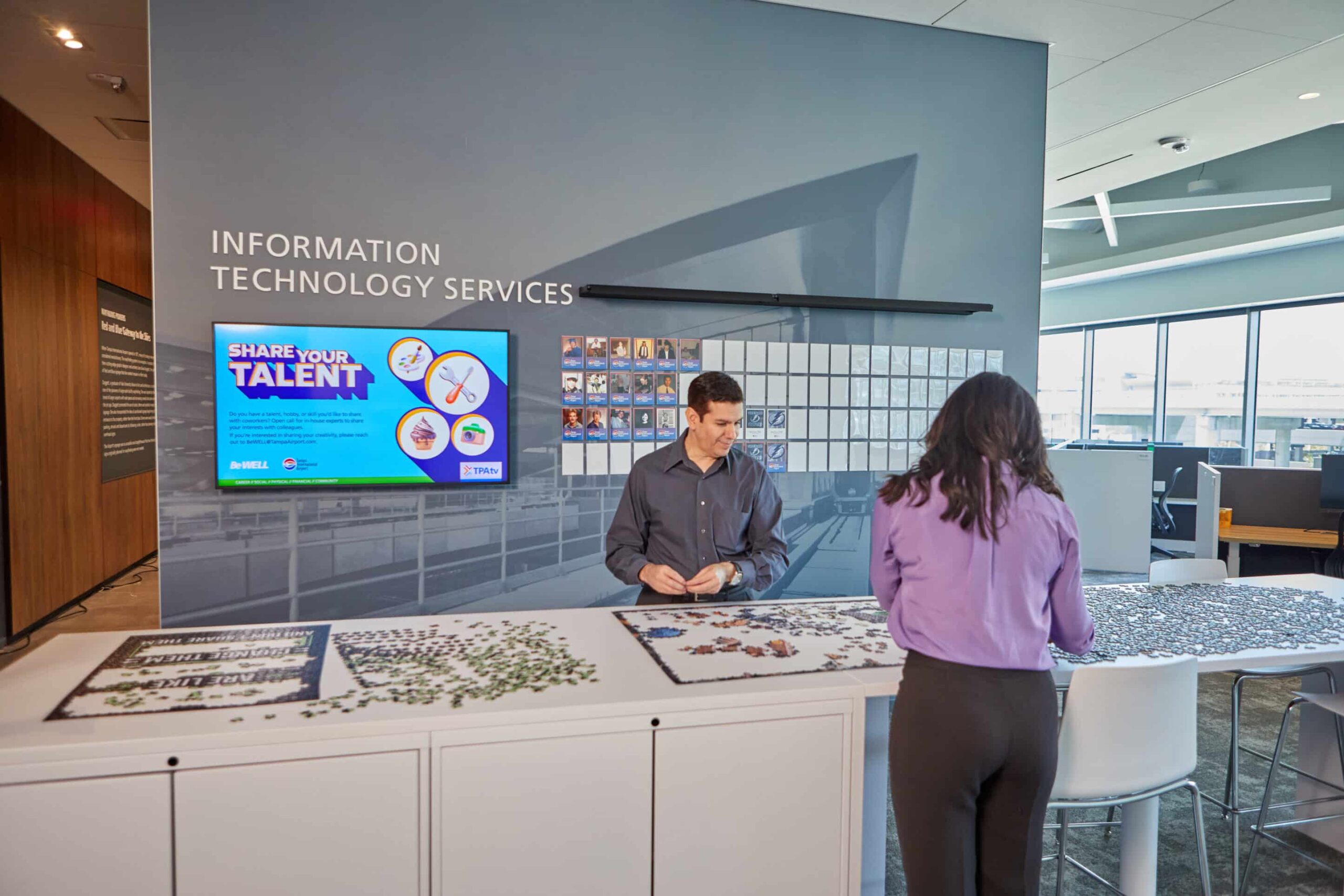 Pair solving puzzles at office, Info Tech Services behind, digital screens/art, natural light through large windows right.