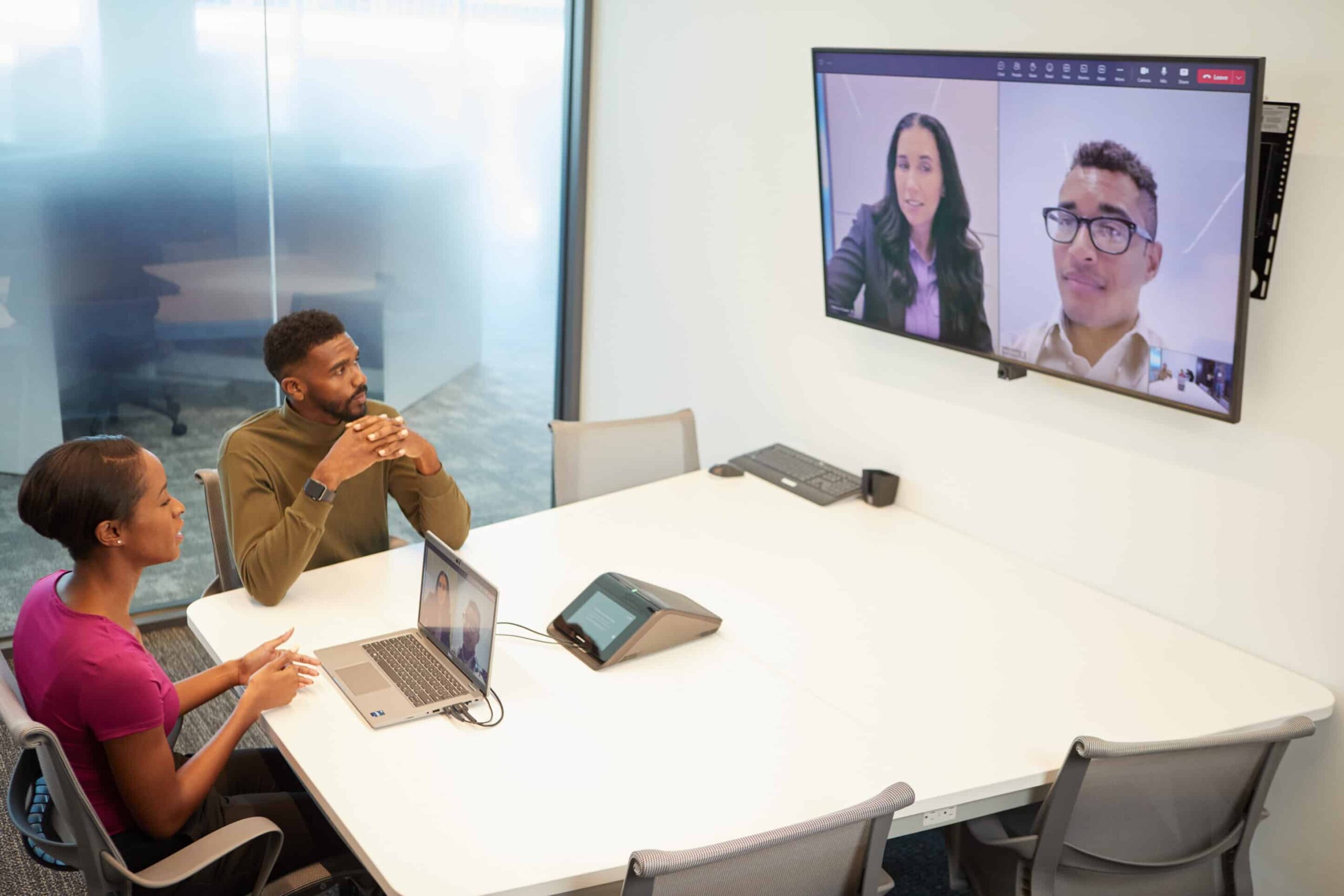 Two people at a conference table on a video call, screen shows two others. Room is bright with large windows.