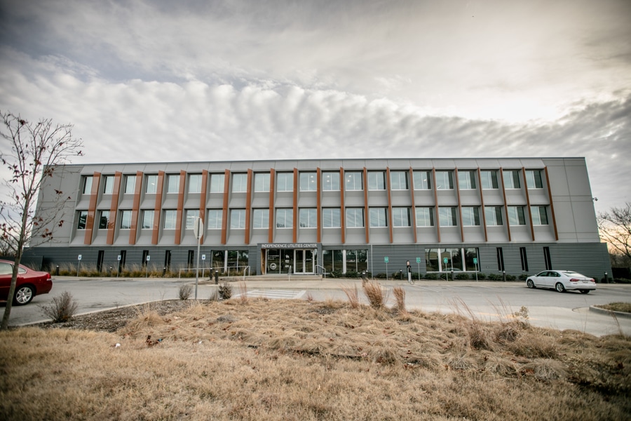 Modern three-story building with large windows, grid facade; cloudy sky, two cars, dry grass, and few trees in the foreground.