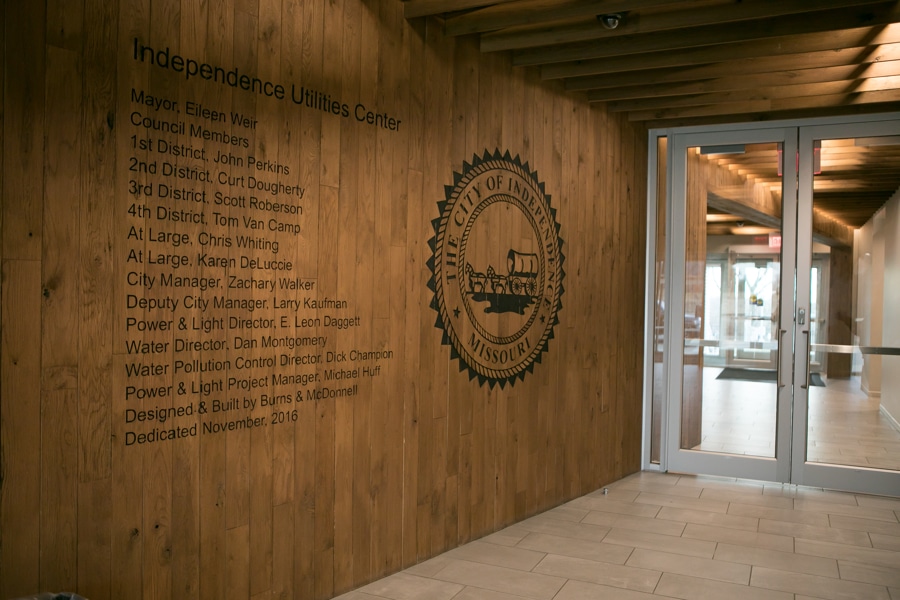 Wooden wall inside building displays city officials list under Independence Utilities Center. Corridor has beige tiles, glass door.