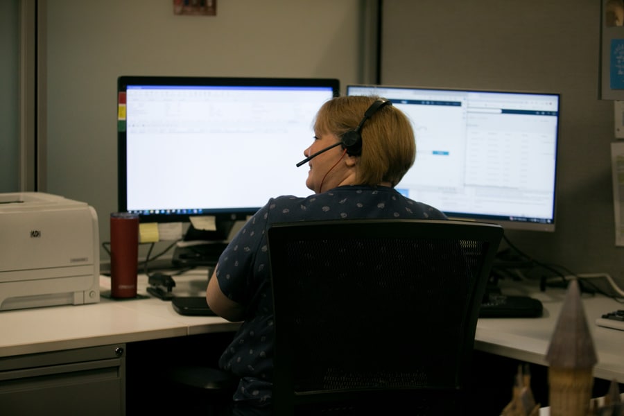 Headset on, person at dual monitors; desk has printer, tumbler, office supplies. Office with partition walls.