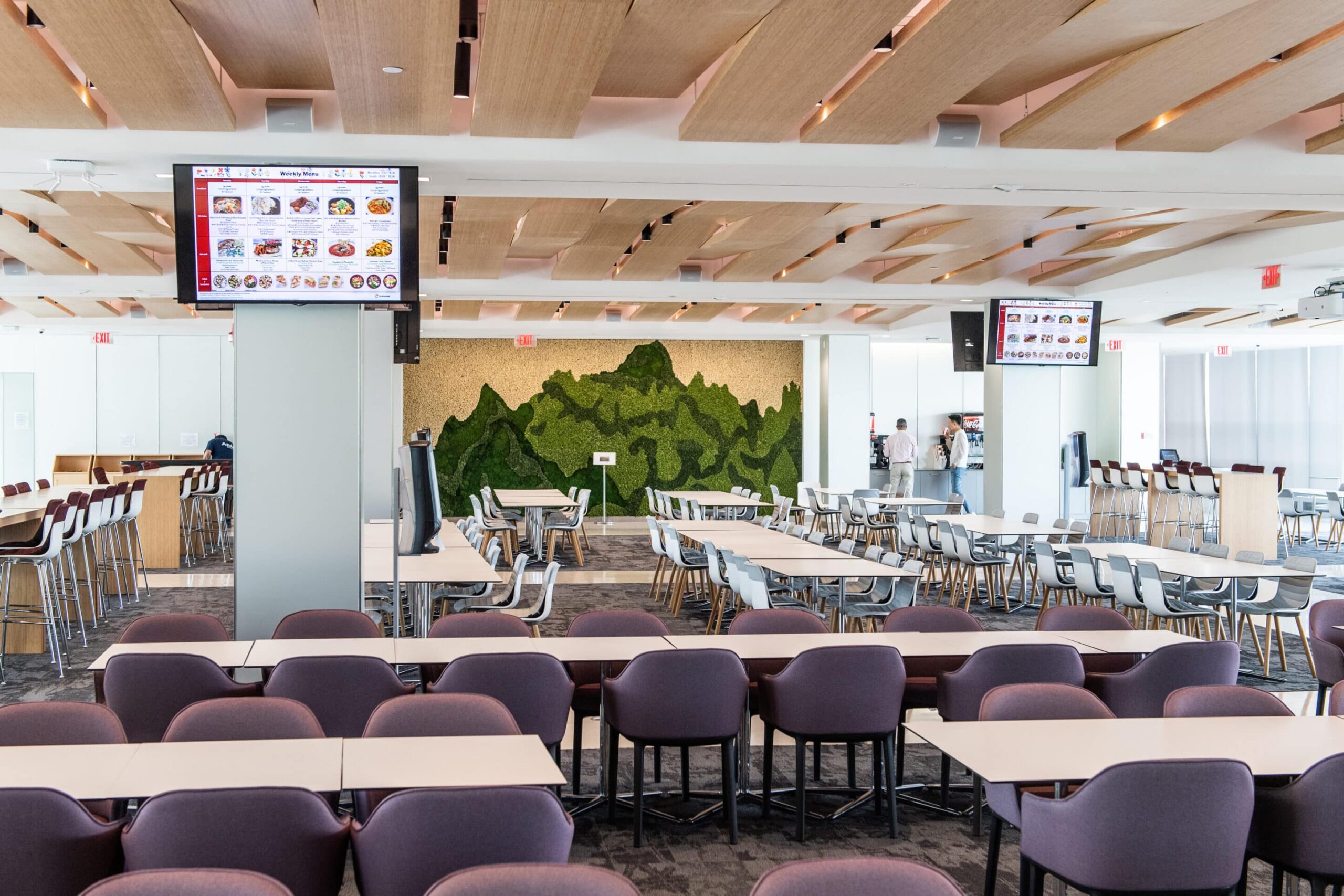 Spacious cafeteria with tables, purple chairs, menu screens, green mountain wall art, wooden ceiling panels, and large windows.