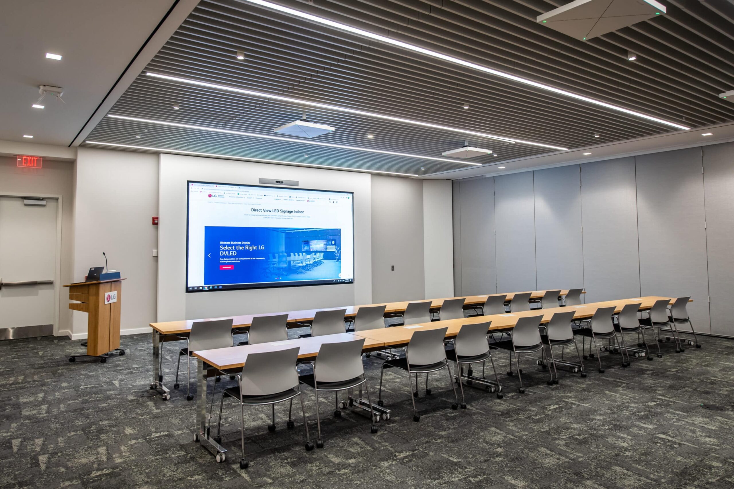 Modern conference room with presentation screen showing a homepage, rows of tables, white chairs, podium near exit.