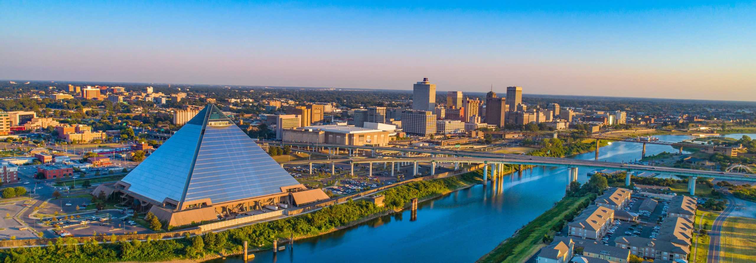 Aerial view of Memphis: pyramid by the river, skyline & bridge in background, residential areas in foreground, clear blue sky.