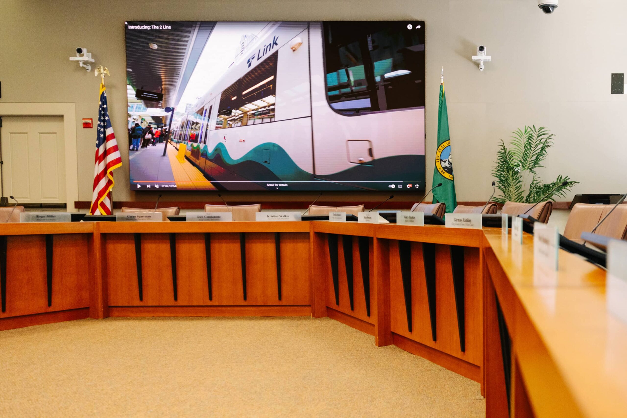 Room A: large screen with paused train video, U-shaped table with nameplates, US flag left, green flag right, plant in corner.
