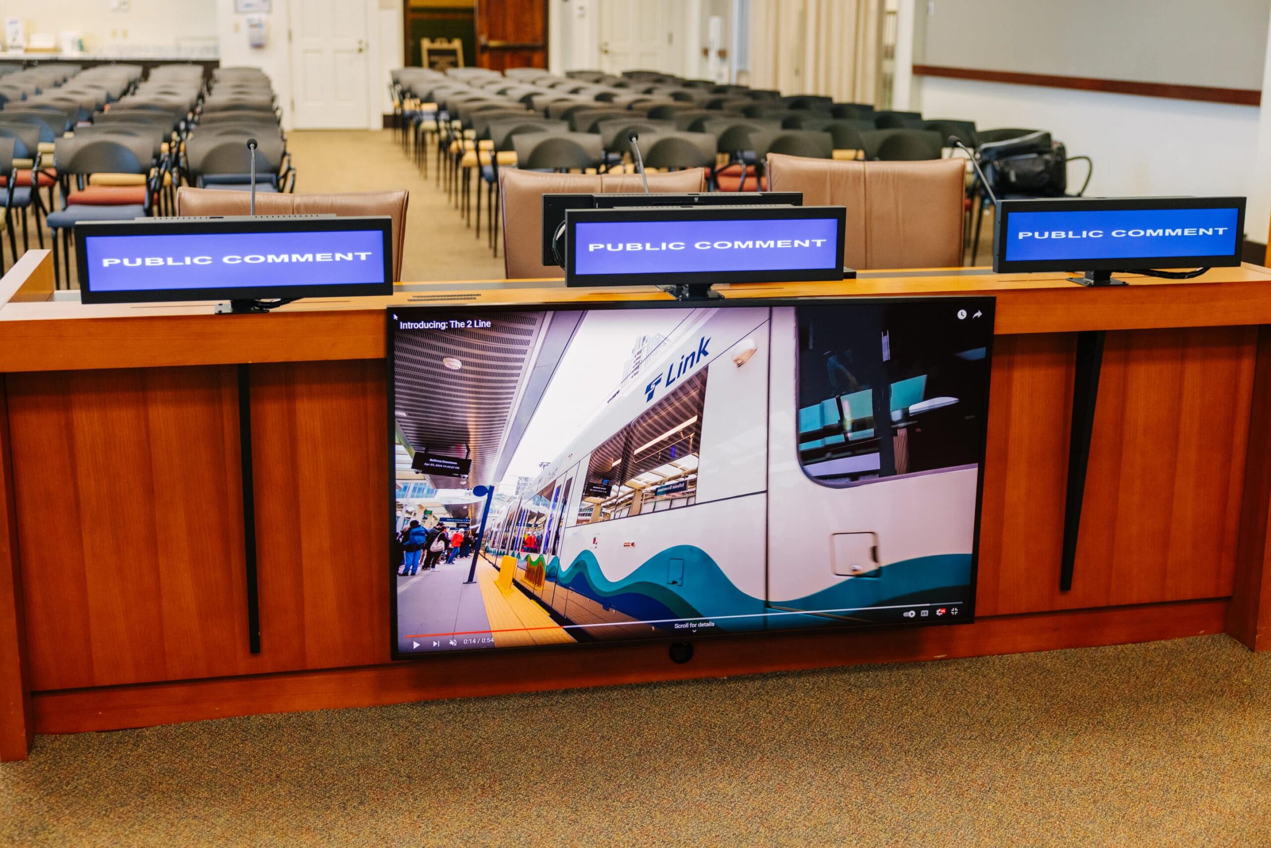 Three Public Comment screens on a desk; below, a large screen shows a train and people. Empty chairs are in the background.