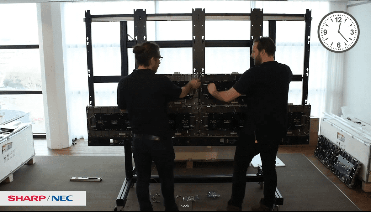 Two in black shirts work on a screen; clock shows 10:10. Sharp/NEC logo, tools, and boxes around. Room with large windows.